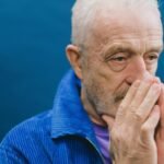 Senior unshaven gray haired male in blue jacket standing against blue background with hands clasped near face while looking away
