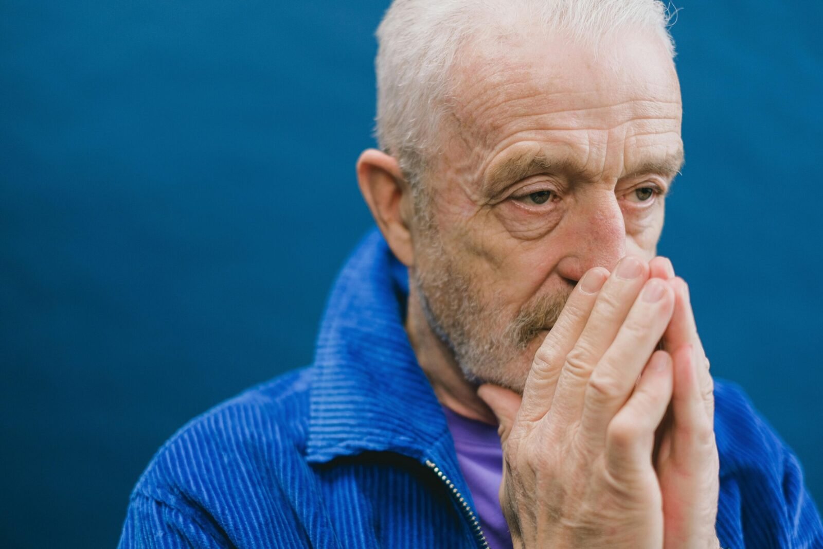 Senior unshaven gray haired male in blue jacket standing against blue background with hands clasped near face while looking away