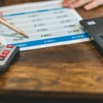 Close-up of a hand with pen analyzing financial rates on paper with a calculator and laptop nearby.