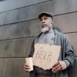 An elderly homeless man holds a cardboard sign asking for help in an urban setting.