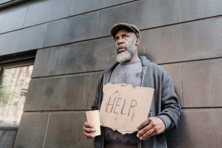 An elderly homeless man holds a cardboard sign asking for help in an urban setting.